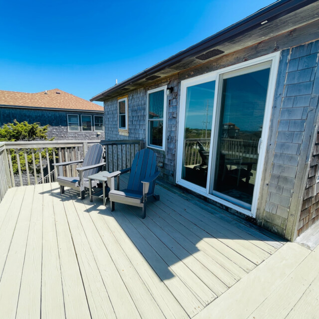 Two Adirondack chairs on a wooden deck outside a shingled house with sliding glass doors under a clear blue sky.
