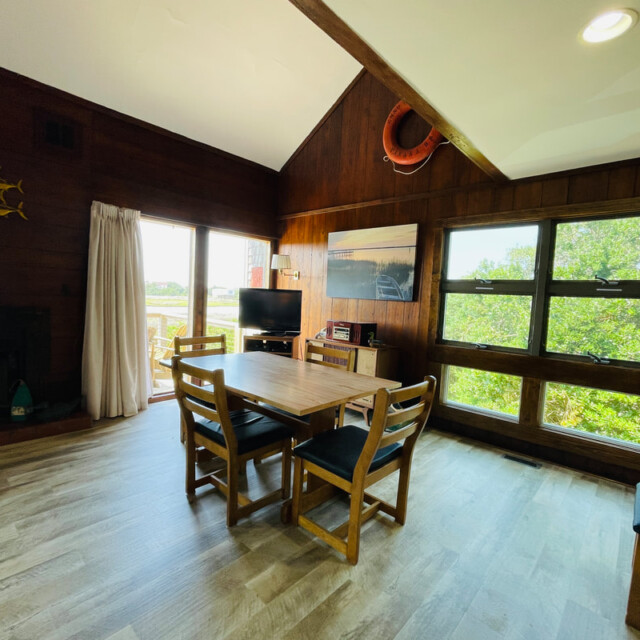 Dining area with wooden table and chairs, TV, artwork, and large windows showing greenery outside.