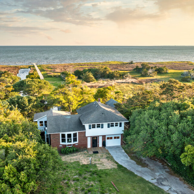 A two-story house surrounded by trees with a view of the coast and a dock leading to the water under a partly cloudy sky.