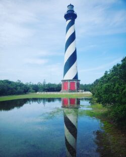 Tall lighthouse with black-and-white spiral stripes reflected in a calm pond, surrounded by greenery and a blue sky.