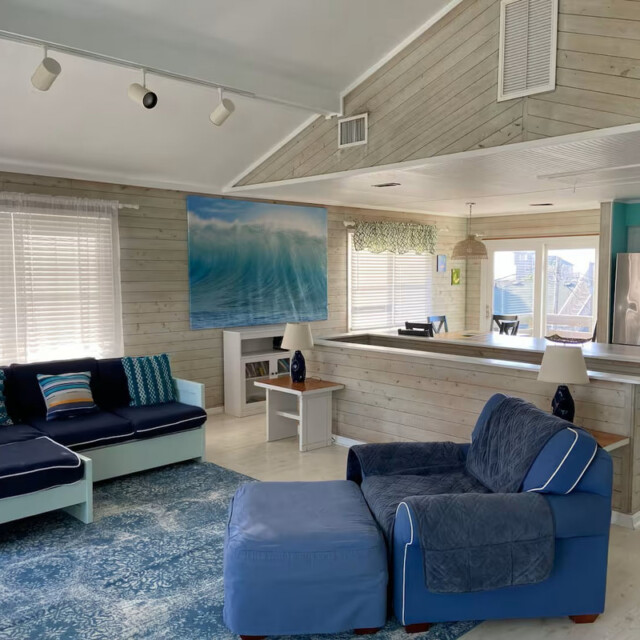Beach-themed living room with blue seating, ocean artwork, and a view into a modern kitchen with stainless steel appliances.