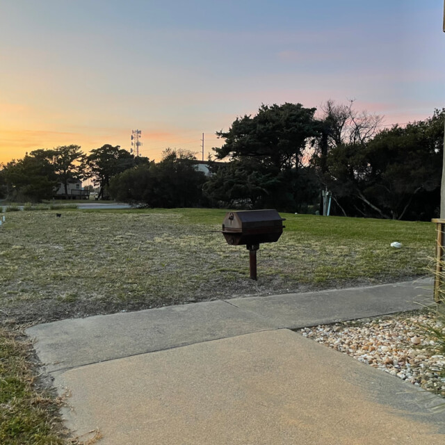 A small charcoal grill stands next to a concrete walkway in a grassy yard at sunset.