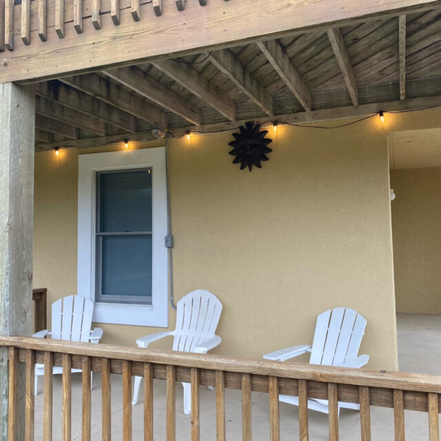 Three white Adirondack chairs sit on a covered porch with string lights and a sun-shaped wall decoration.