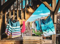 Swimwear and towels hang on a clothesline under wooden stairs with a bright blue sky in the background.