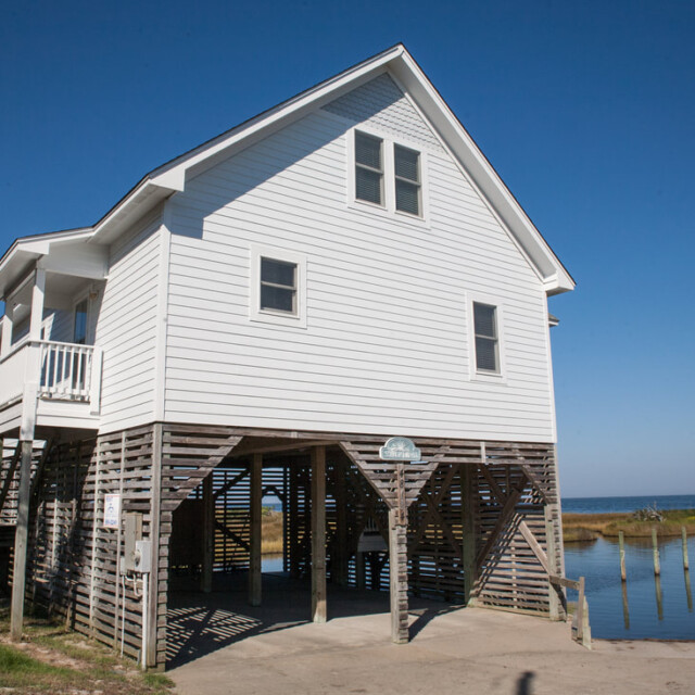 A white stilt house stands near the water, with a clear blue sky and dock visible in the background.