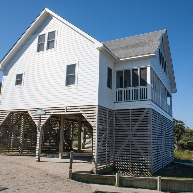 White elevated house with lattice skirting, screened porch, and sloped roof by water on a clear, sunny day.
