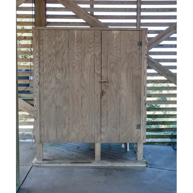 A wooden cabinet with double doors and a latch stands on a concrete floor in front of a slatted wooden wall.