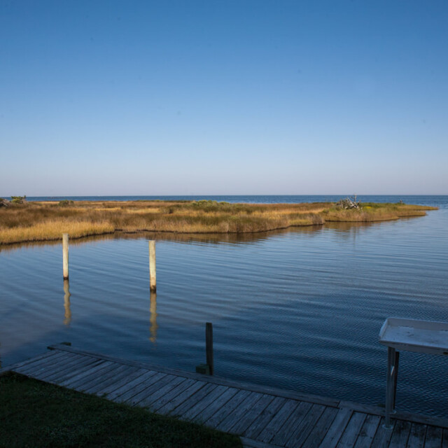 Wooden dock extends over calm water with marsh grasses and clear blue sky in the background.