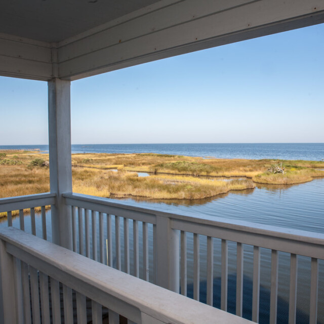 View from a covered wooden deck overlooking marshland leading to a calm body of water under a clear blue sky.