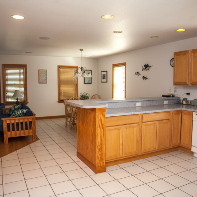 Open kitchen with wooden cabinets, white tile floor, and adjacent living area with blue sofa and dining table.
