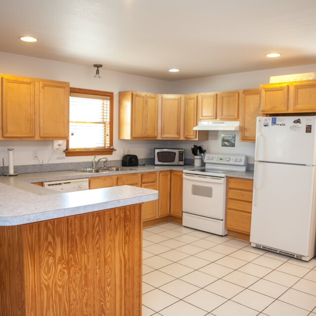 Bright kitchen with light wood cabinets, white appliances, tile floor, and a breakfast bar with three wooden stools.