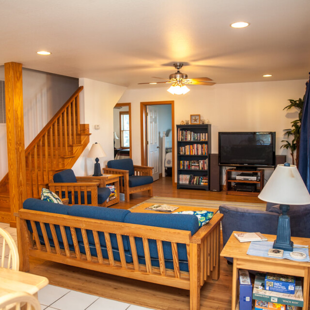 Living room with wooden furniture, blue cushions, a TV, bookshelves, board games, and a wooden staircase.