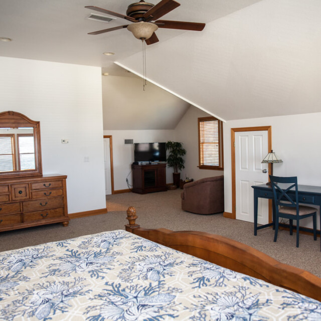 Spacious bedroom with wooden furniture, a blue desk, ceiling fan, TV, and patterned bedspread under sloped ceiling.