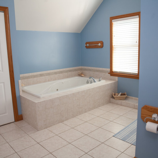 Bathroom with a large bathtub, blue walls, tiled floor, window with blinds, and a wall-mounted toilet paper holder.