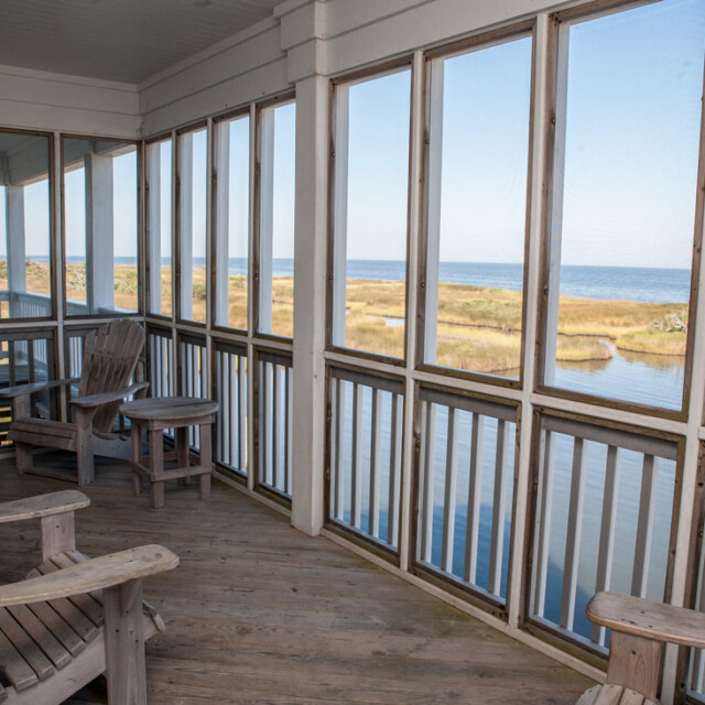 Screened porch with wooden chairs overlooks marshland and water under a clear sky.