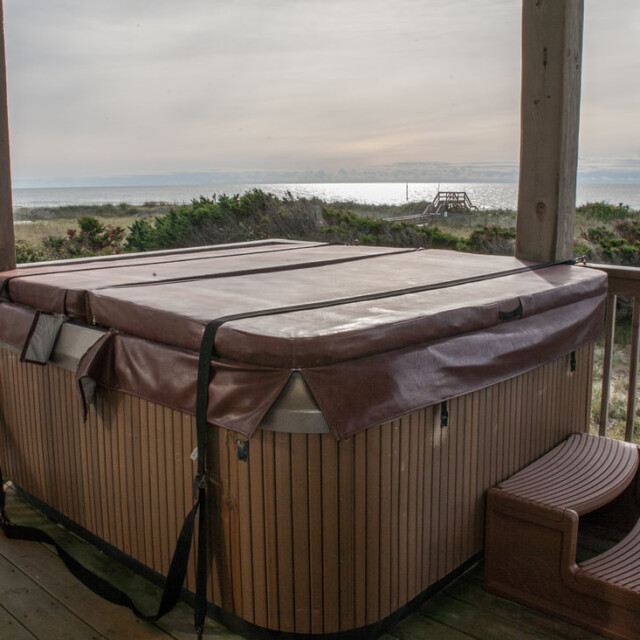 A closed hot tub sits on a wooden deck overlooking a beach and ocean under an overcast sky.