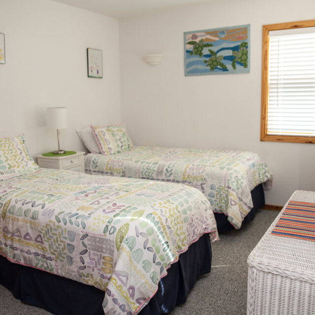 Bedroom with two twin beds, pastel bedding, a window with blinds, wicker furniture, and colorful wall art.