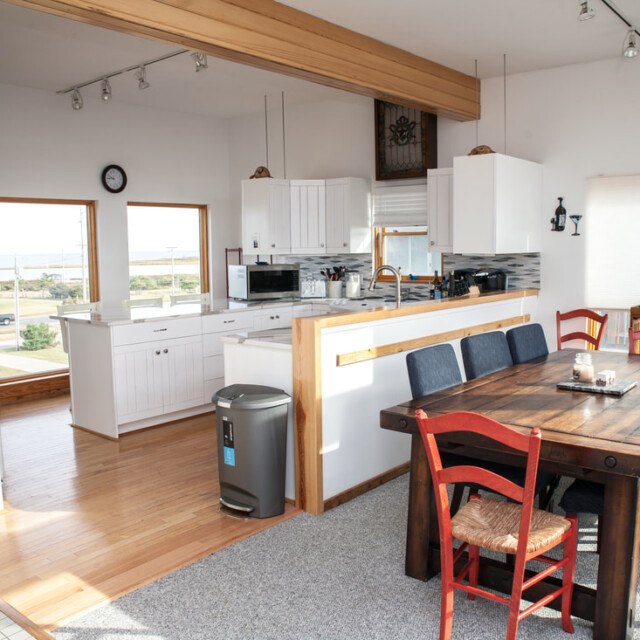 Open kitchen and dining area with white cabinets, wooden dining table, mixed chairs, and large window with outdoor view.