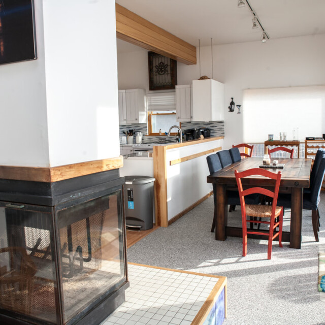 Open kitchen and dining area with a wooden table, mismatched chairs, fireplace, and large windows letting in sunlight.