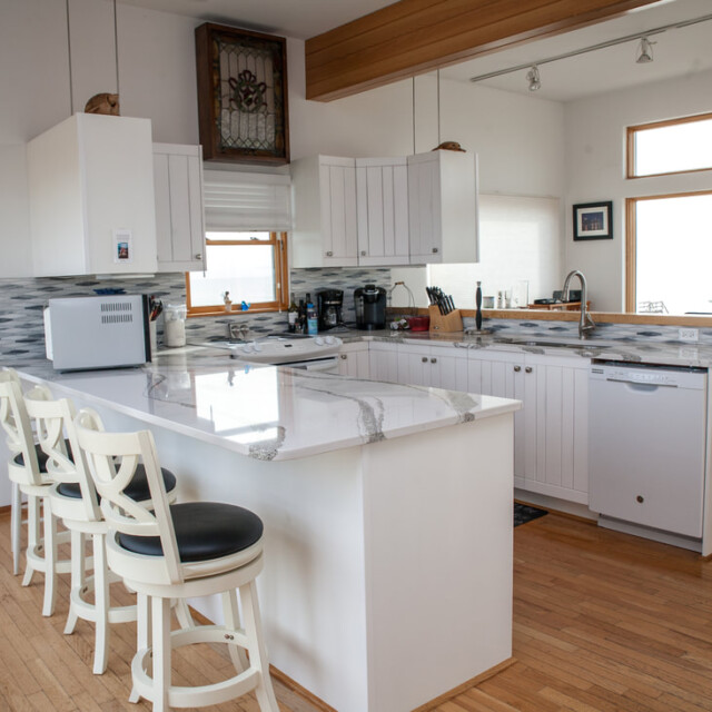 Modern kitchen with white cabinets, marble-patterned countertops, three barstools, and wood flooring.
