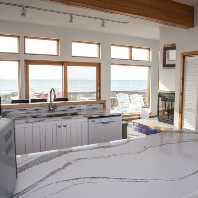 Modern kitchen with white cabinets, marble countertop, large windows, and an ocean view in the background.