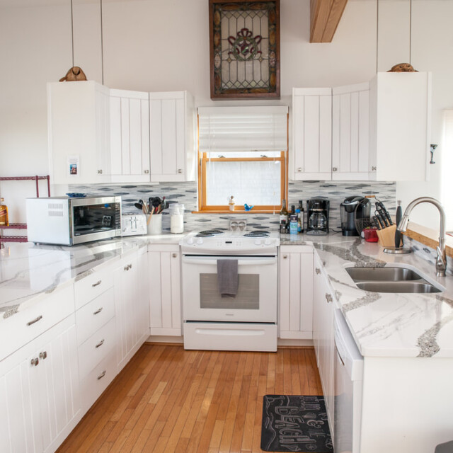 Modern white kitchen with marble countertops, electric stove, microwave, coffee maker, sink, and wooden floors.
