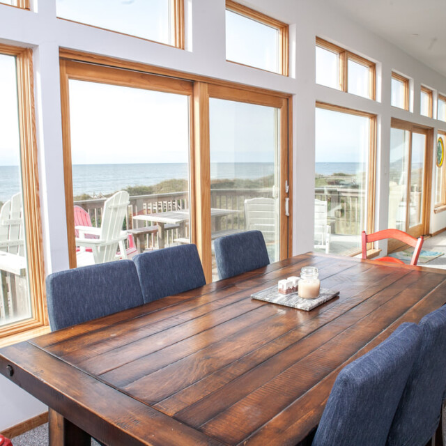 Wood dining table and blue chairs in a bright room with large windows overlooking a beach and ocean view.