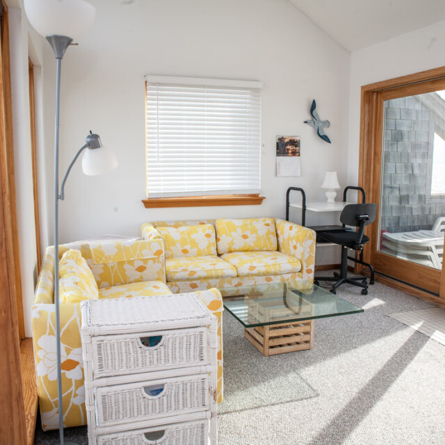 Small sunlit living room with a yellow floral sofa, glass coffee table, desk, and sliding doors to a patio.