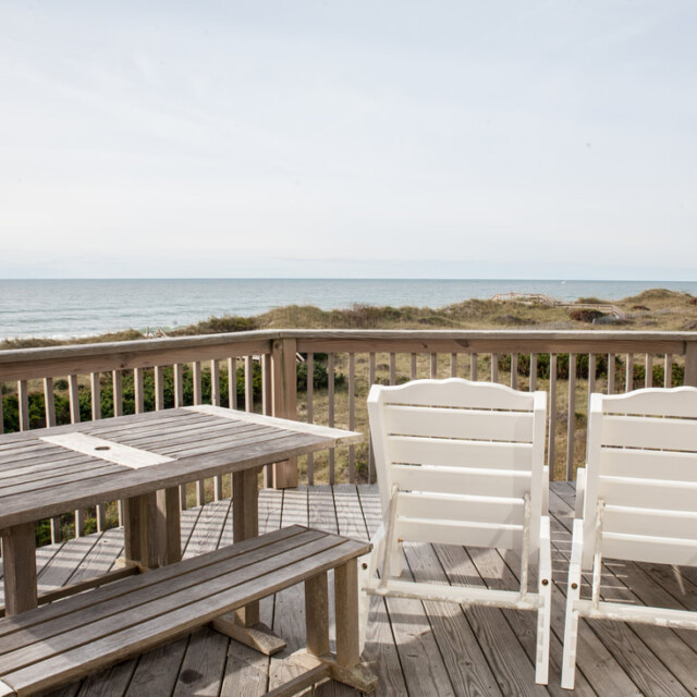 Wooden deck with two white chairs and a table overlooks sand dunes and the ocean under a clear sky.