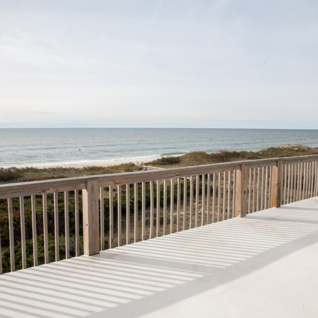 View of a wooden deck overlooking sand dunes and the ocean under a partly cloudy sky.