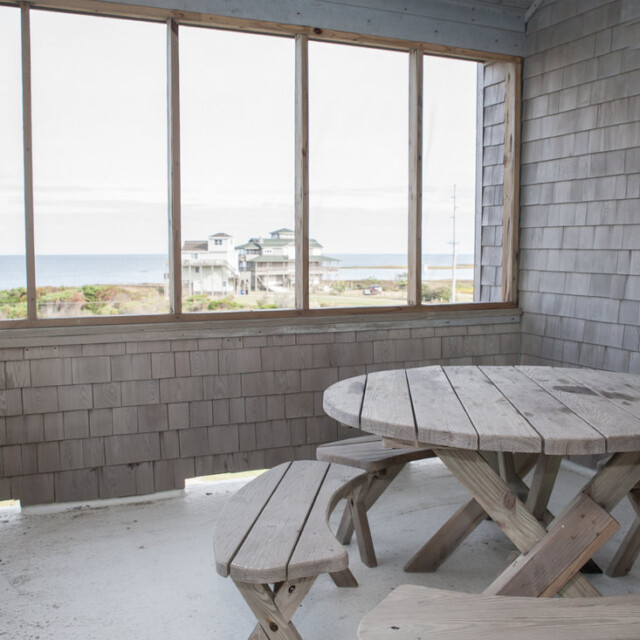 Covered patio with a round wooden picnic table and benches, overlooking the ocean and nearby houses through screened windows.