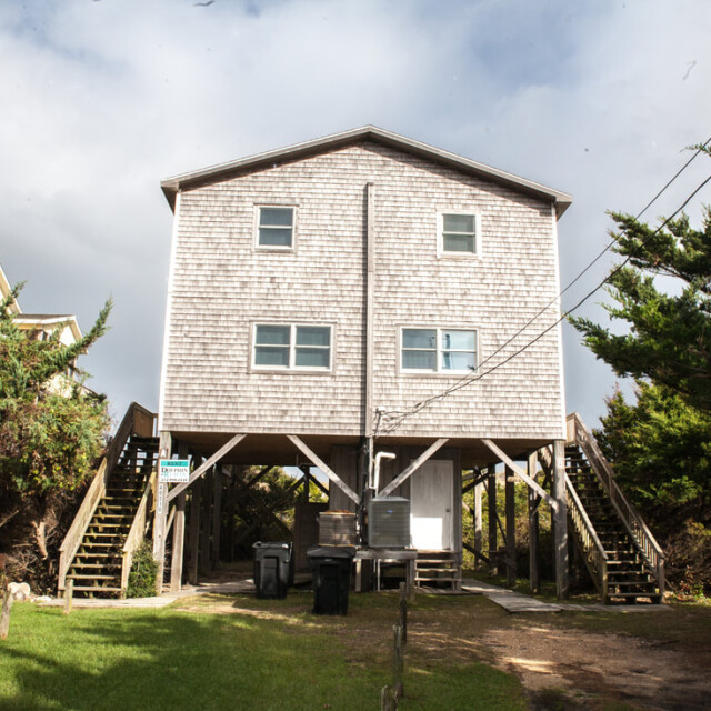 Two-story wooden beach house on stilts, surrounded by trees, with stairs on both sides and a grassy yard in front.