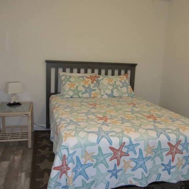 A small bedroom with a double bed, starfish-patterned bedding, a nightstand, lamp, and light walls.