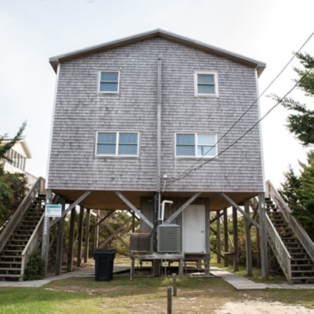 Two-story wooden house on stilts with stairs on both sides, surrounded by trees and a grassy yard.