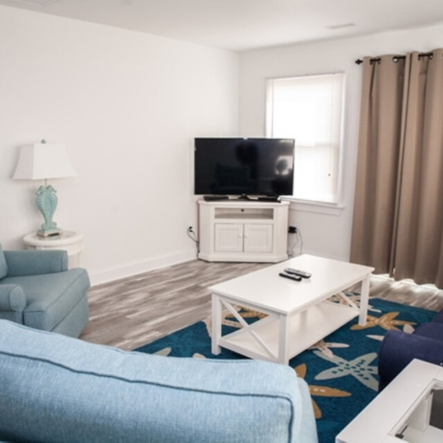 Living room with blue chairs, a TV on a stand, white coffee table, lamp, and beige curtains on the window.