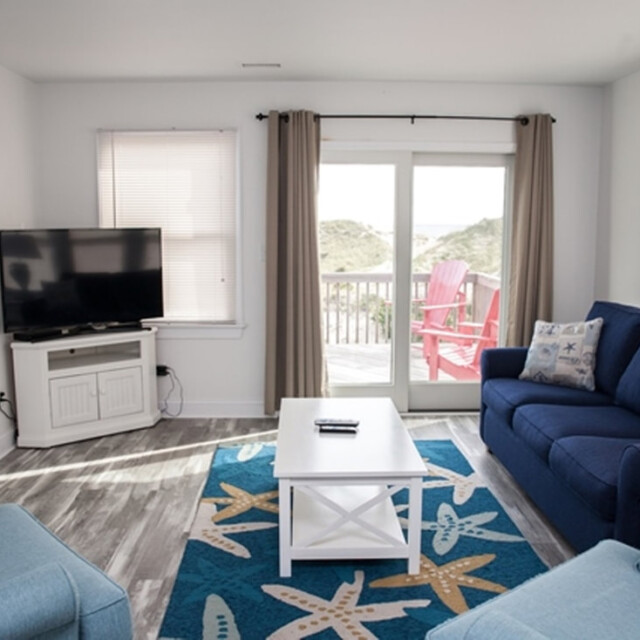 Living room with blue furniture, a TV, a white coffee table, and glass doors leading to a deck with pink chairs outside.