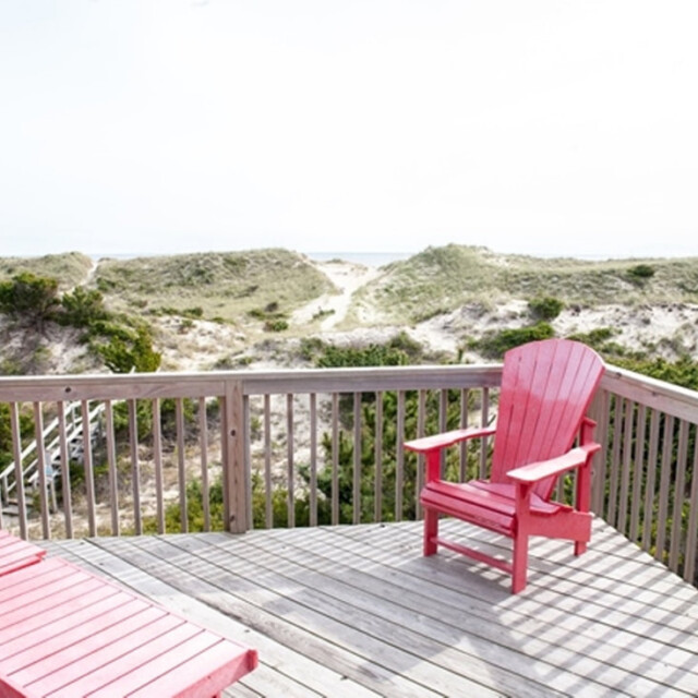 Wooden deck with red Adirondack chairs overlooks sandy dunes and greenery under a bright, clear sky.